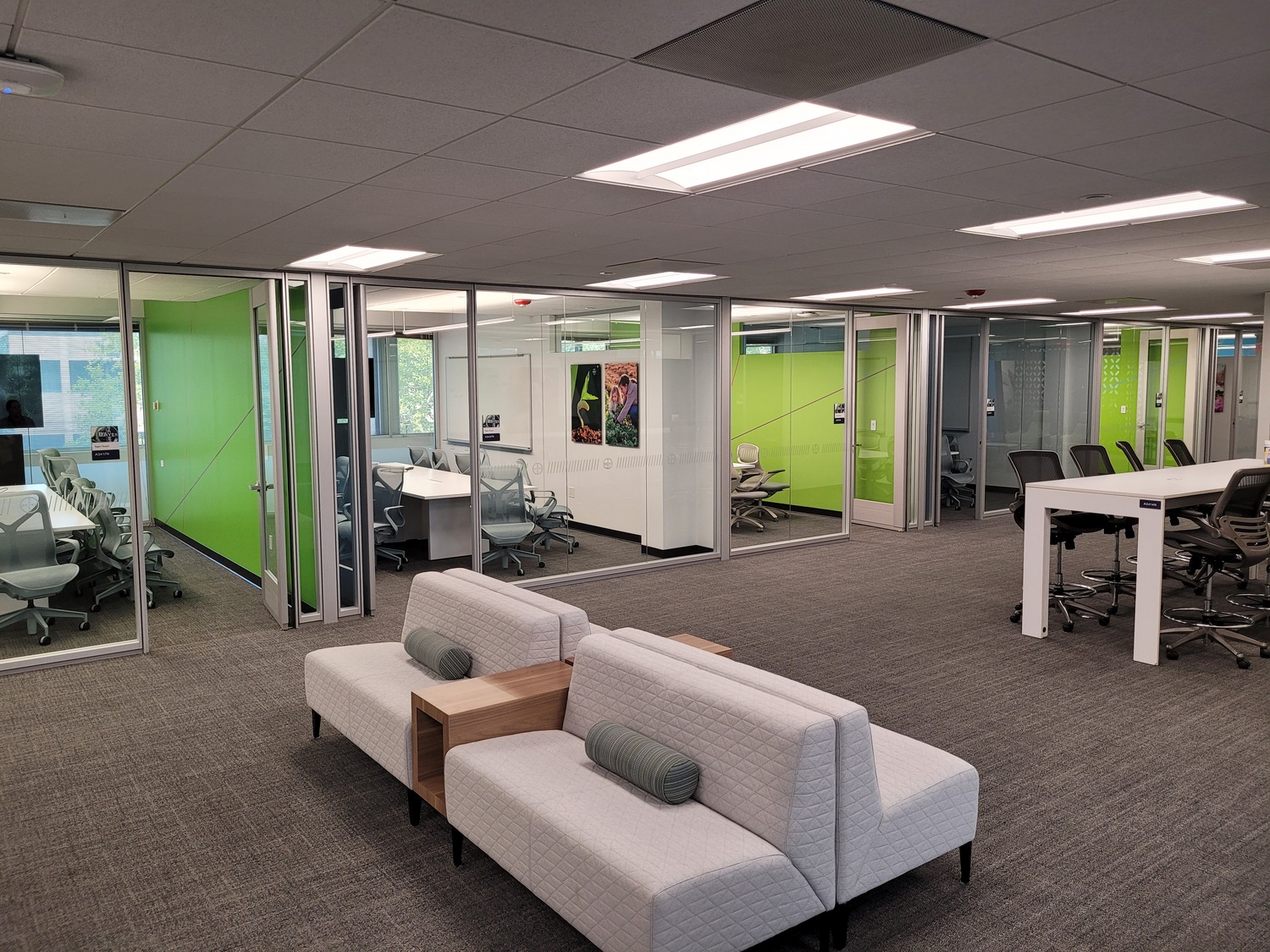 Modern office lobby at Bayer A3 North featuring a collaborative seating area with back-to-back beige couches and wooden accent tables. The space includes six glass-walled conference rooms with sleek furniture and writable surfaces, complemented by a contemporary meeting table with ergonomic chairs. The design showcases Horner & Shifrin’s approach to creating functional, aesthetically pleasing corporate environments.
